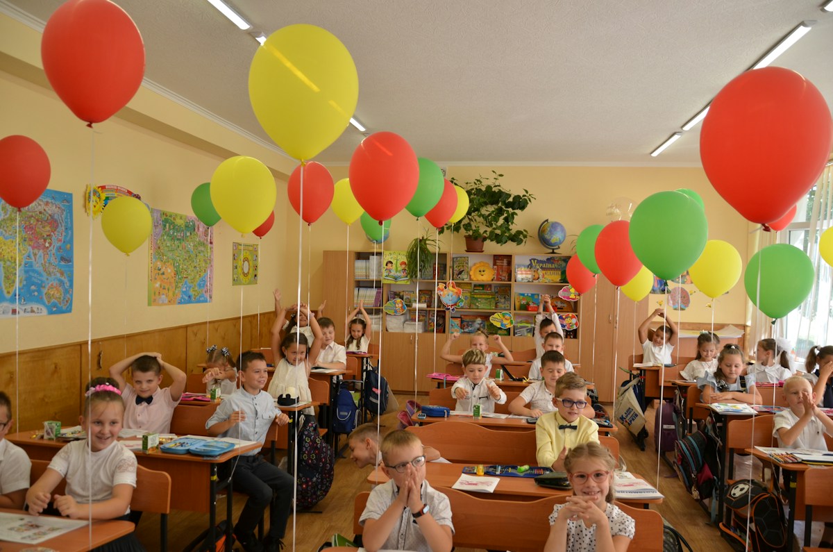 Illustrative photo: a bright classroom of smiling schoolchildren at their desks, with red, yellow and green balloons tied to every desk