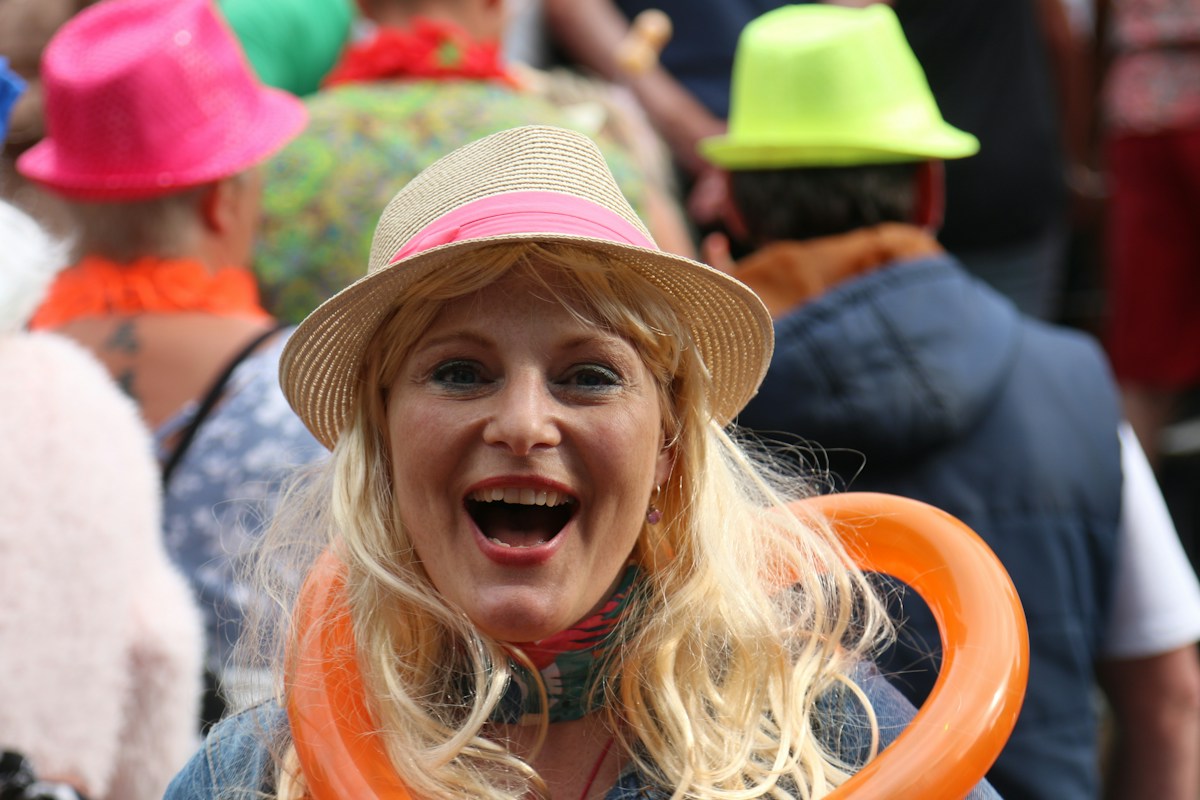 Illustrative photo: a laughing woman in a straw hat at a street festival, wearing an orange twisted balloon loop around her neck