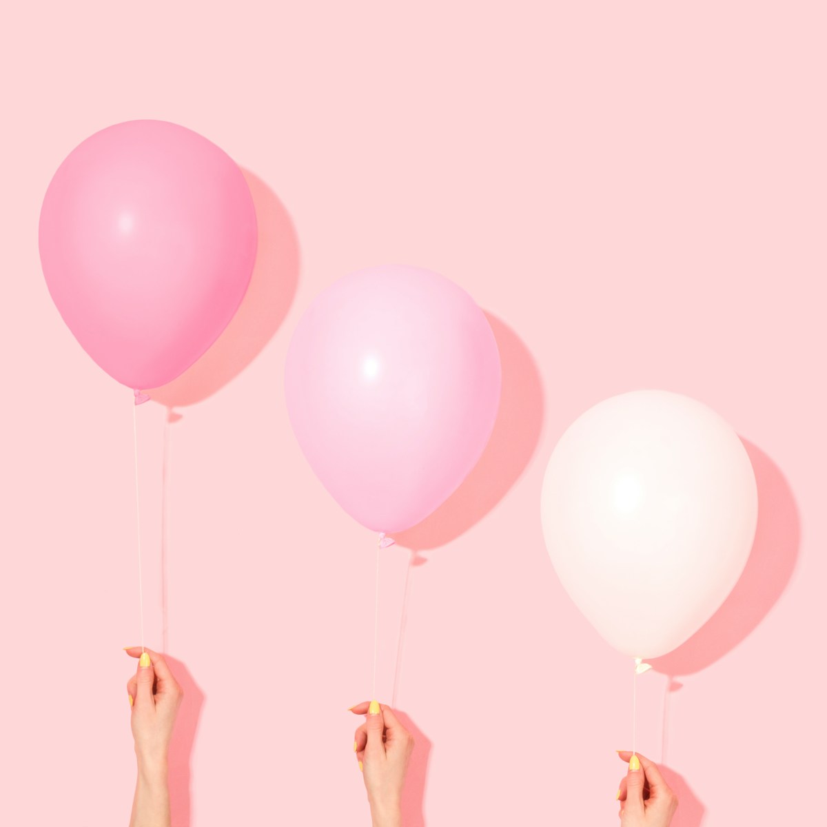 Illustrative photo: three hands holding up three balloons in hot pink, light pink and white against a blush-pink wall