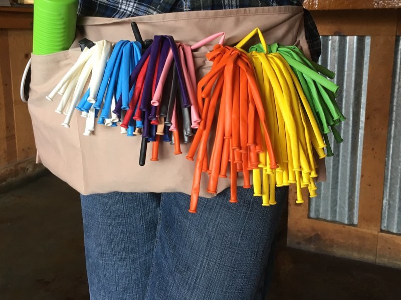 Illustrative photo: a balloon twister's canvas apron with pockets full of uninflated long balloons in every colour, and a green hand pump
